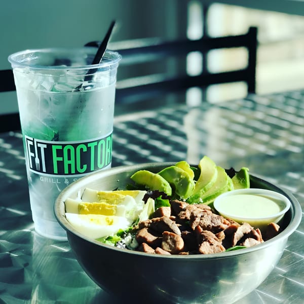 A Fit Factory Grill salad with chicken, avocado, and egg, next to a branded cup of water.