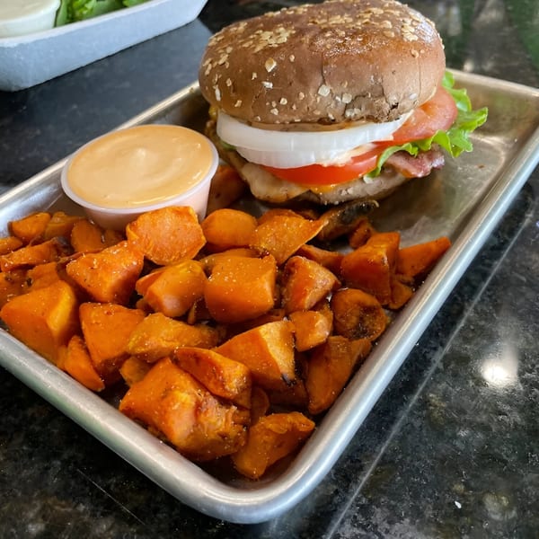 A Fit Factory Grill burger with sweet potato fries and dipping sauce on a metal tray.