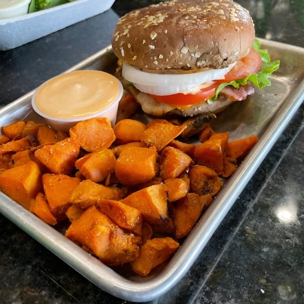 A Fit Factory Grill burger with sweet potato fries and dipping sauce on a metal tray.