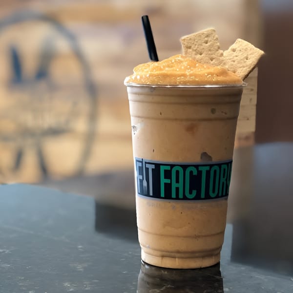 A Fit Factory Grill smoothie with a straw and two graham crackers on a dark counter.